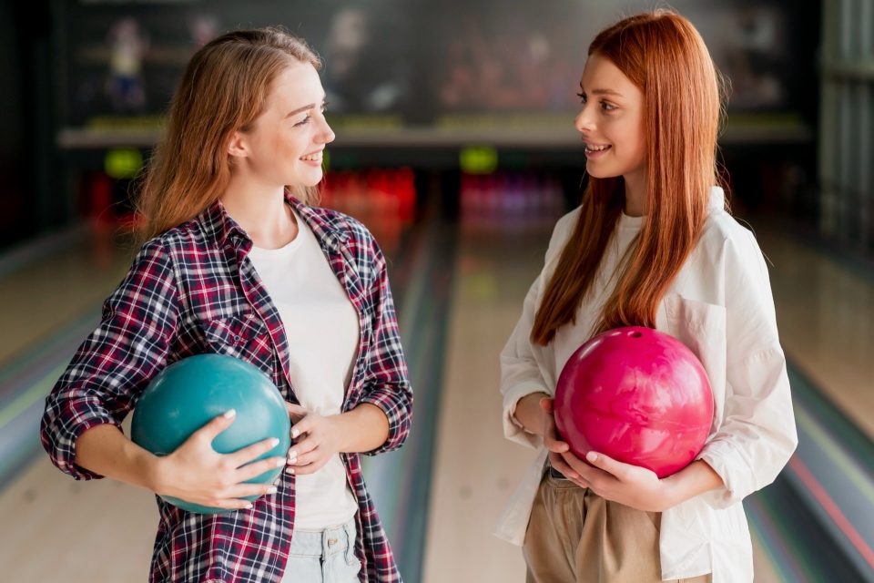 Två leende kvinnor håller bowlingklot och ler mot varandra i en bowlinghall